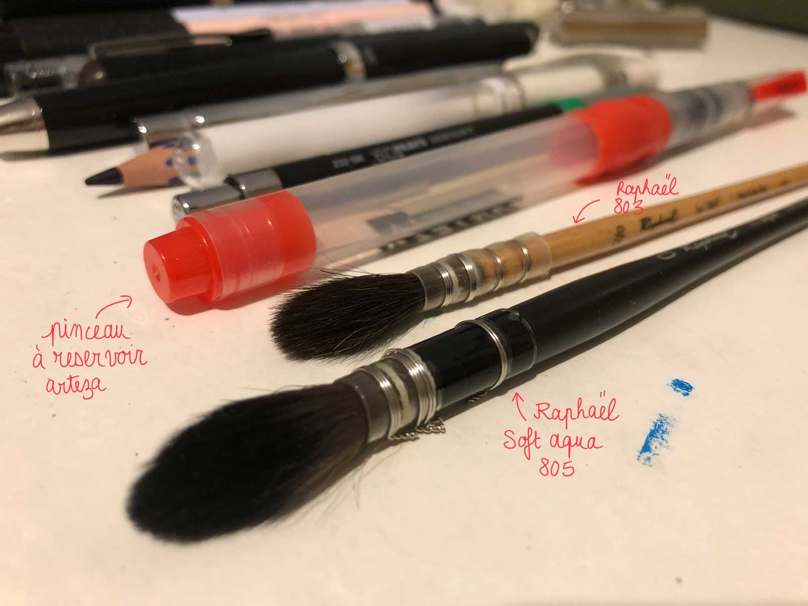 on voit deux pinceaux à lavis, le pinceau à réservoir à eau et les autres stylos et crayons en arrière plan.